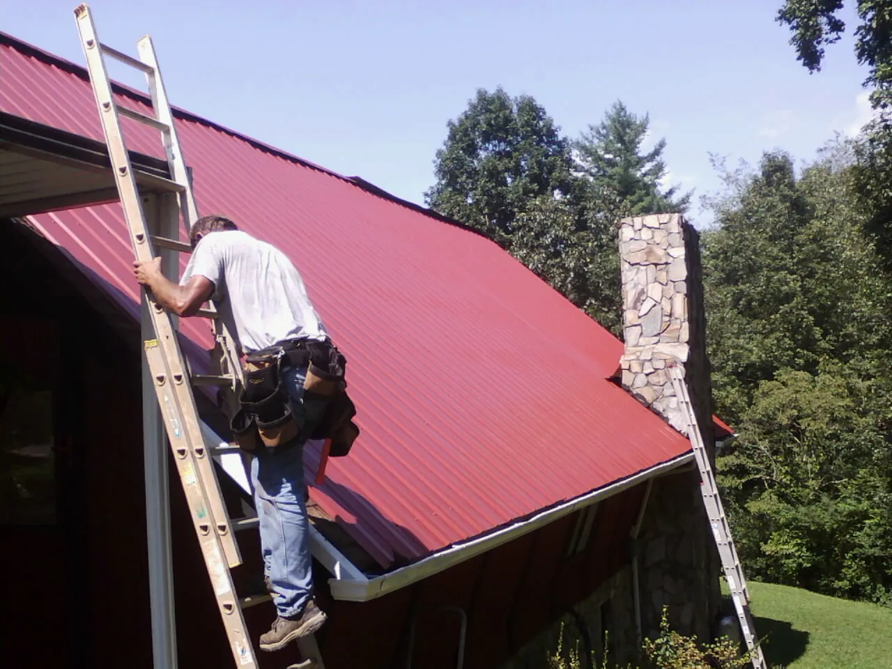 Roofer on a ladder beside a red metal roof and stone chimney — Soffit & Fascia Repair by Roofing Brothers in Westpoint